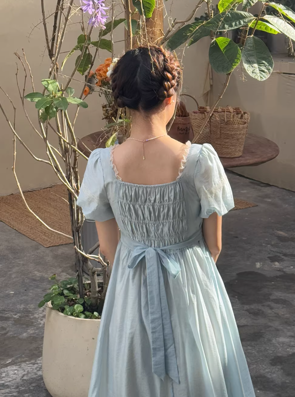 Woman in a princesscore light blue dress with white rose embroidery sitting on a vintage wooden bench, embodying a dreamy fairytale aesthetic.