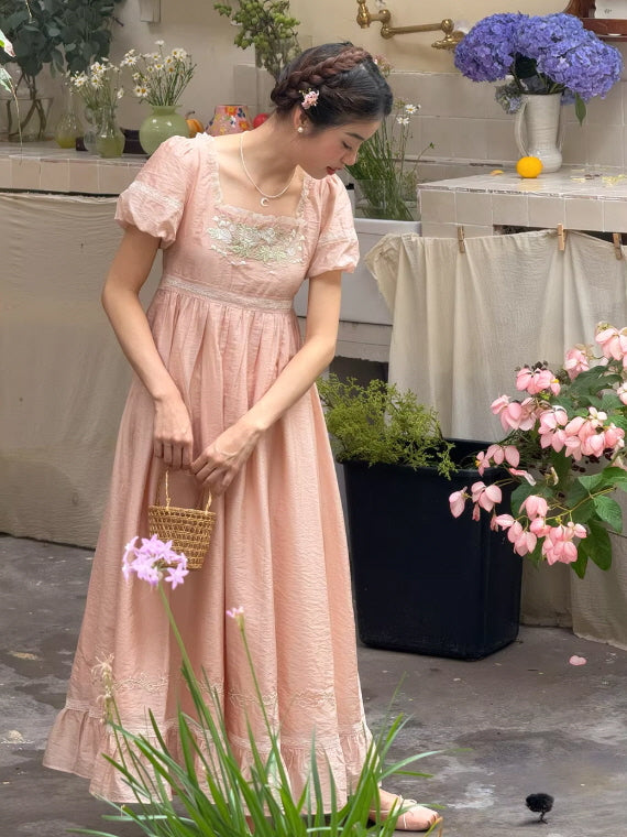 Romantic pink cottagecore dress with puff sleeves and empire waist, woman holding a small wicker basket in a fairytale garden setting with hydrangea flowers.
