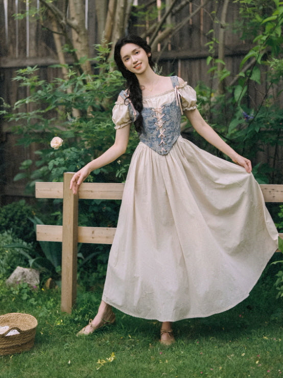 Blue floral corset cottagecore maxi dress with puff sleeves and a flowing beige skirt in a romantic garden setting