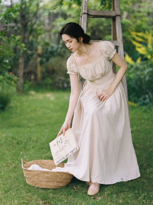 Beige prairie maxi dress with puff sleeves and gathered waist styled with a woven basket in a cottage garden