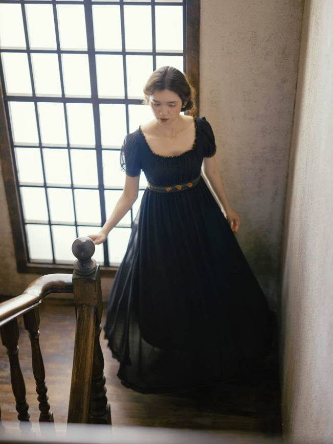Elegant black regency inspired maxi dress with empire waist and flowing skirt beside a vintage staircase window