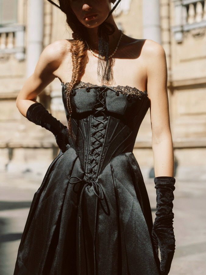Full-length black vintage corset dress with a lace-up front and flowing midi skirt, styled with a wide-brim hat and long gloves.
