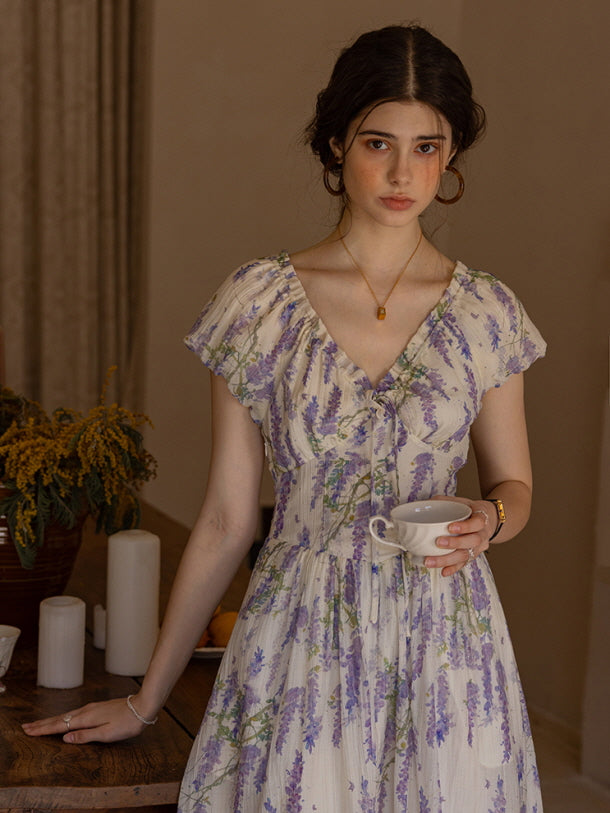 Young woman in a lavender floral dress, holding a delicate teacup while gazing softly at the camera, exuding a calm and vintage-inspired aesthetic with warm, muted tones.