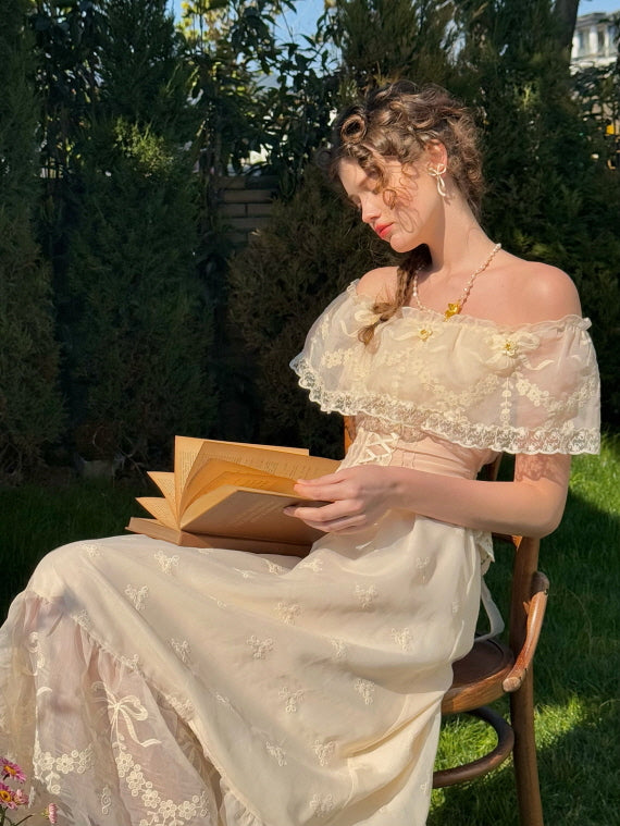 Woman in ivory off-shoulder lace corset dress reading a vintage book in a sunlit garden, featuring floral embroidery and romantic fairytale style