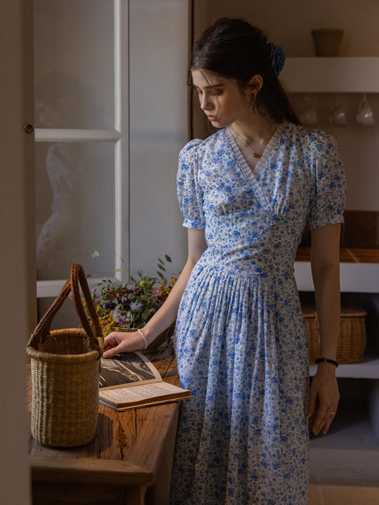 A woman wearing a blue floral print dress with puff sleeves, standing by a wooden table with a woven basket and an open book, exuding a serene cottagecore vibe in a rustic indoor setting.