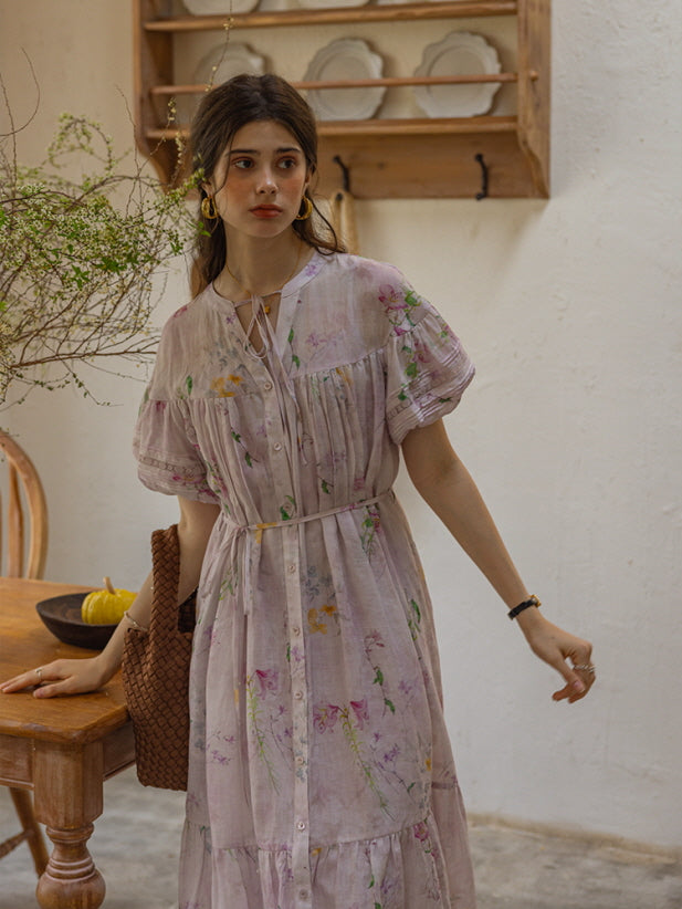 A young woman stands beside a rustic kitchen counter, wearing a flowing lilac floral print dress with puff sleeves and a delicate lace-up neckline, exuding a dreamy, cottagecore vibe.