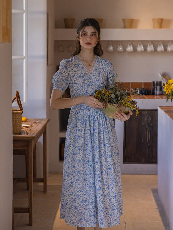 A woman in a vintage-inspired blue floral dress holding a rustic flower arrangement in a ceramic bowl, set against a cozy kitchen background with warm, muted tones and classic decor.
