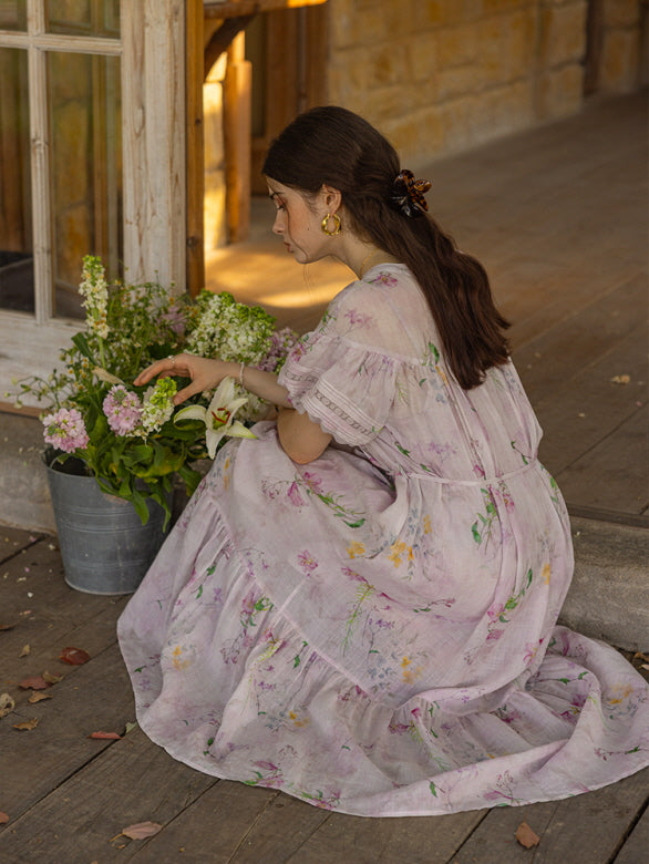 A woman in a lilac floral tiered dress kneels beside a bucket of fresh flowers, her hair adorned with a claw clip, creating a serene and romantic cottagecore aesthetic.
