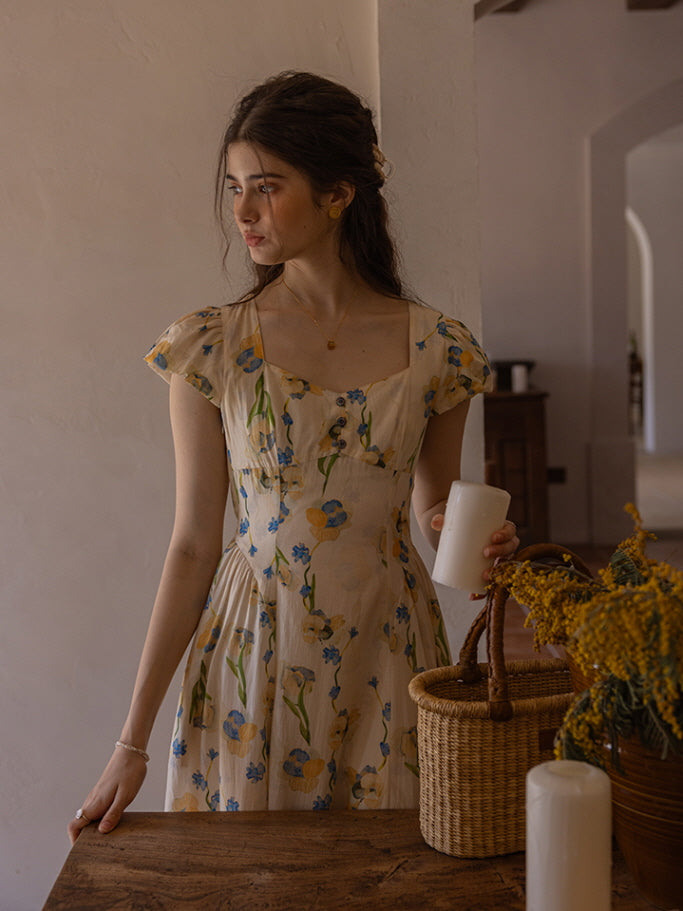 A young woman with soft, wavy hair gazes thoughtfully while holding a white candle. She wears a cream floral dress adorned with blue and yellow blossoms, creating a romantic, vintage-inspired ambiance in a softly lit room with a woven basket and dried flowers.