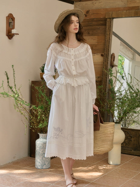 A woman wearing a white vintage-style blouse and skirt set with delicate ruffle and lace details, accessorized with a straw hat and straw bag, standing in a cottagecore-inspired interior garden setting.