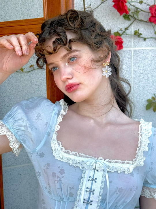 A close-up shot of a model wearing a pastel blue floral corset top with lace trim and puff sleeves, adorned with daisy-shaped earrings, evoking a soft, romantic feel against a backdrop of red roses.