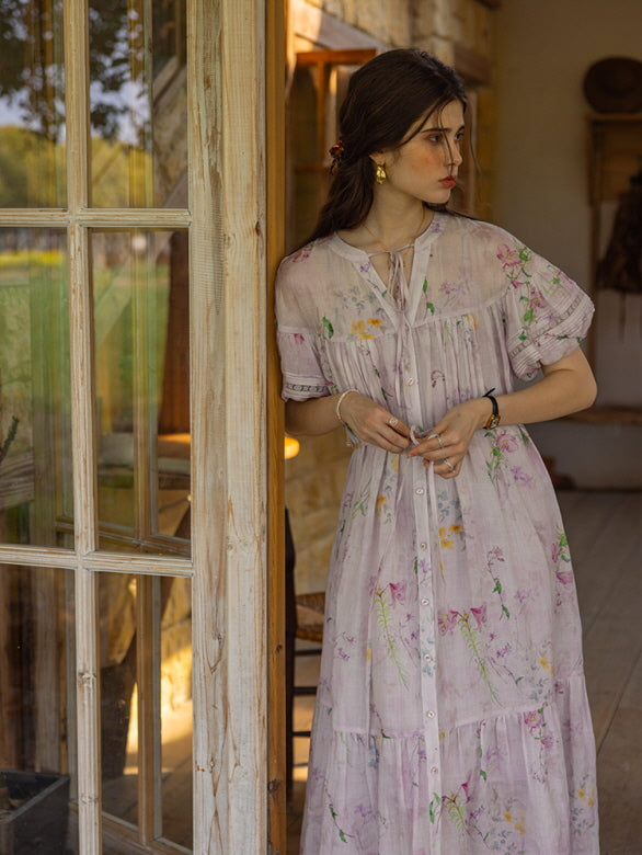 The model sits on a wooden floor surrounded by pastel floral arrangements, wearing a flowy, vintage-inspired dress with delicate botanical prints and a gathered waist.