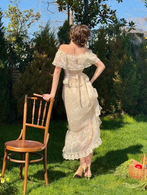 Back view of a woman in an ivory lace corset dress with ribbon tie and ruffle hem, standing in a garden under sunlight – vintage romantic fairytale fashion
