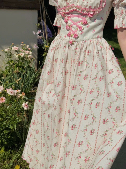 Close-up of a soft ivory cotton dress with delicate pink rose pattern, corset-style ribbon detail and gathered waist, exuding a vintage cottagecore aesthetic.