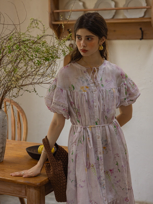 A woman stands near a wooden kitchen counter, wearing a soft floral print midi dress with puff sleeves and a waist tie, paired with a brown woven bag. The natural light illuminates her gentle expression.