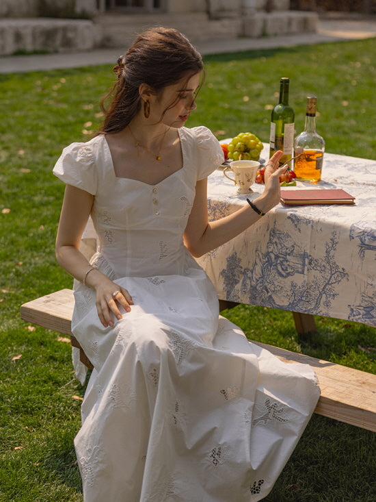 A woman seated on a wooden bench in a garden, wearing a white embroidered cotton dress with delicate floral lace patterns, holding a book and surrounded by a table setting with fruits and wine bottles. 
