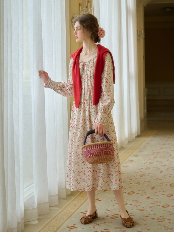 A young woman wearing a cottagecore floral outfit with a red sweater draped over her shoulders, holding a woven basket bag, standing by a sheer white curtain in a bright, vintage-style hallway.