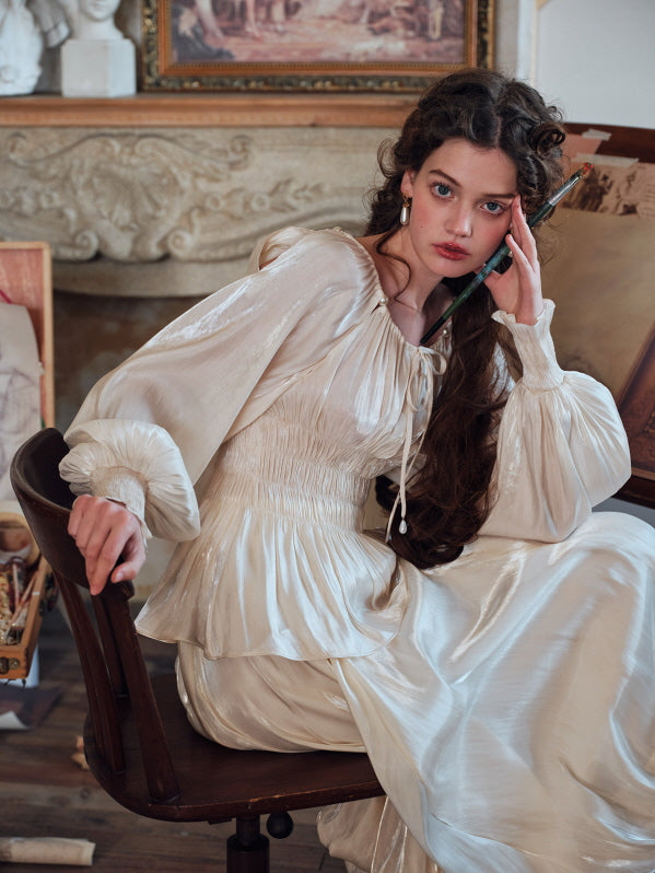 A woman wearing a vintage-inspired ivory blouse with puff sleeves and a ruched waist, standing gracefully in an art studio adorned with classical paintings and wooden easels, embodying a romantic, cottagecore aesthetic.