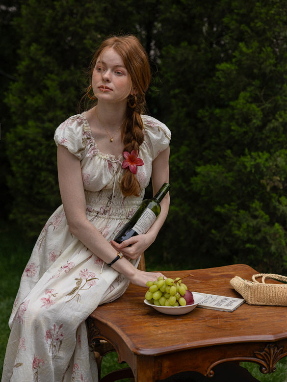 A woman sitting at a vintage wooden table holding a wine bottle, styled in a floral cottagecore dress with ruffled neckline and smocked waist. Romantic, fairytale vibe.
