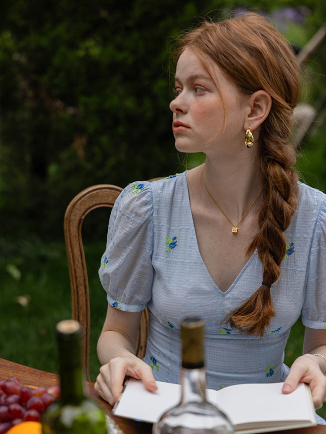 A woman with a braided hairstyle gazing thoughtfully to the side while seated at a wooden table outdoors. She is wearing a delicate light blue floral dress with subtle check patterns and puff sleeves, holding a notebook. 