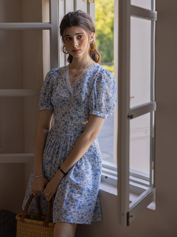 A young woman stands near an open window, wearing a blue floral print short dress with puff sleeves and a delicate lace-trimmed neckline. She holds a woven basket, creating a cottagecore, vintage-inspired look.