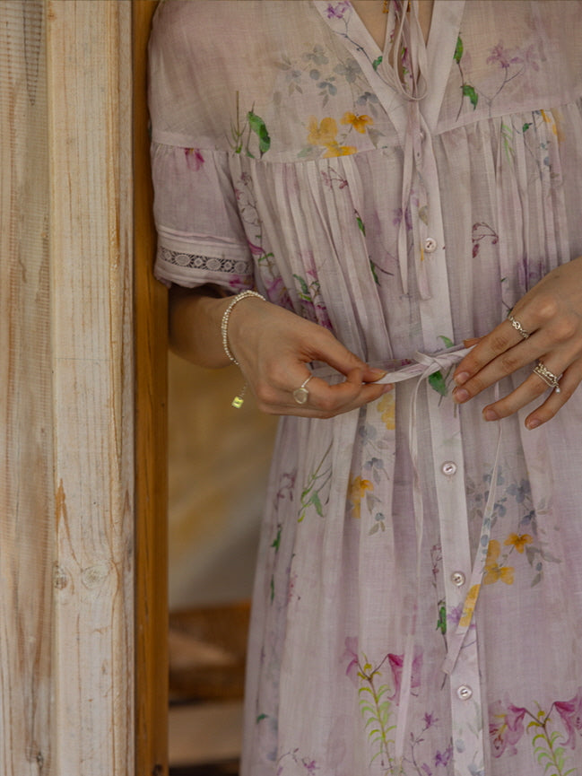 A woman adjusting the waist tie of a romantic lavender floral dress with button-down front, soft gathers, and botanical prints.
