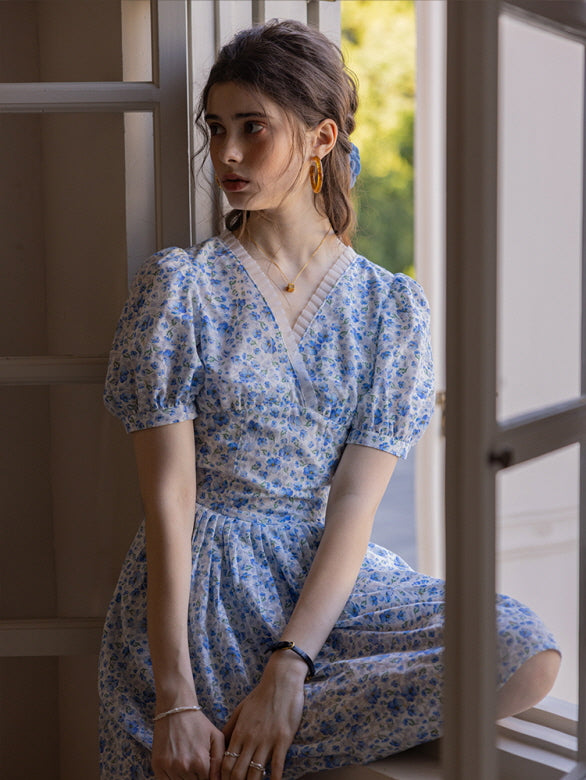 A woman seated by an open window, dressed in a blue floral puff sleeve dress with lace-trimmed neckline. The soft natural light and serene expression evoke a romantic, vintage-inspired mood.