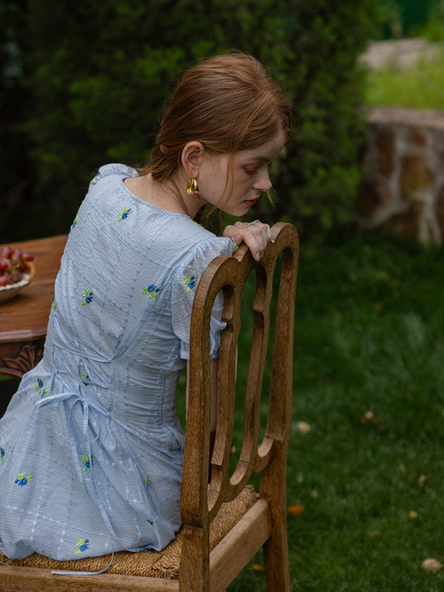 Back view of a woman in a light blue floral dress with a fitted corset-style back, seated on a rustic wooden chair in a lush garden. 