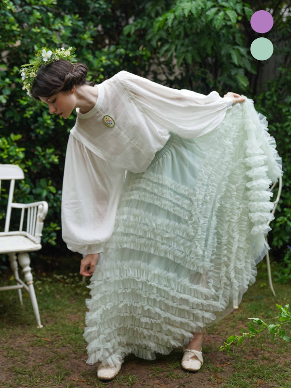 Wearing a voluminous ruffle fairycore skirt and puff-sleeve blouse, the model bends slightly in a dreamy pose, surrounded by lush greenery and flowers.
