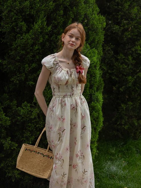 A woman standing against lush greenery, holding a floral embroidered woven bag while wearing a floral cottagecore dress with a smocked waist and ruffled neckline.