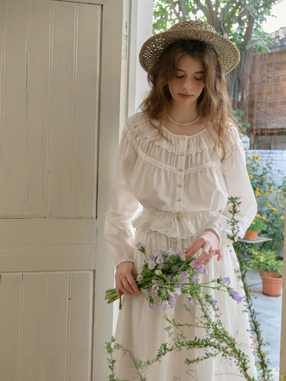 A young woman wearing a white vintage-inspired cottagecore blouse and skirt, holding a bouquet of purple flowers while standing by a wooden door, with a natural and aesthetic atmosphere.