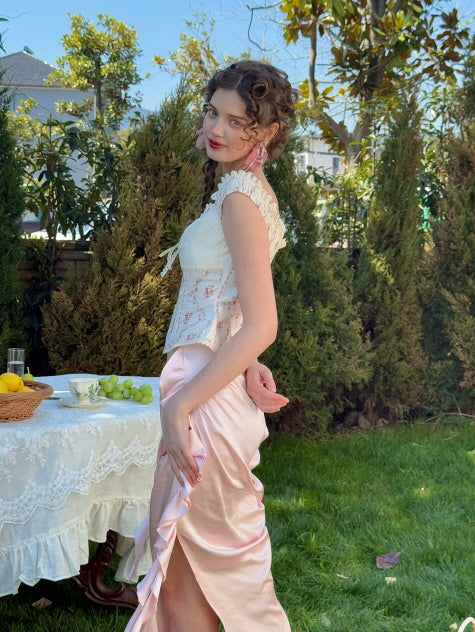 Side view of pastel pink ruched satin skirt with ribbon detail, paired with floral off-shoulder corset top – romantic fairytale-inspired women’s outfit in garden setting