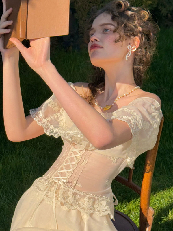 Romantic woman in ivory lace corset dress reading a book under the sun, featuring floral embroidery and ruffled off-shoulder neckline – vintage fairytale fashion aesthetic