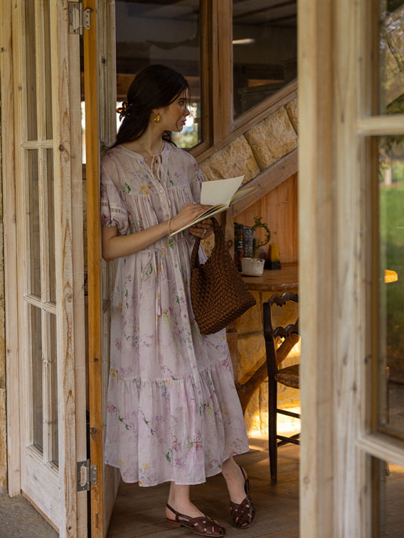 A woman in a soft, floral-printed, lilac midi dress with puff sleeves, standing by a wooden door while holding a book and a woven bag. The rustic, cottage-style background enhances the dreamy, fairytale aesthetic.