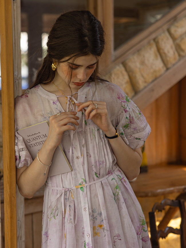 Close-up of a woman in a pastel floral dress, delicately tying the neckline ribbon while holding a book titled "Quantum Statistics." The soft, vintage-inspired setting creates a nostalgic, poetic mood.