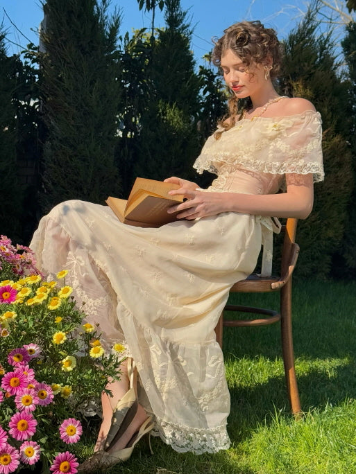 Romantic woman in ivory lace corset dress reading a book on a wooden chair beside blooming garden flowers – vintage fairytale aesthetic with ruffle and embroidery details