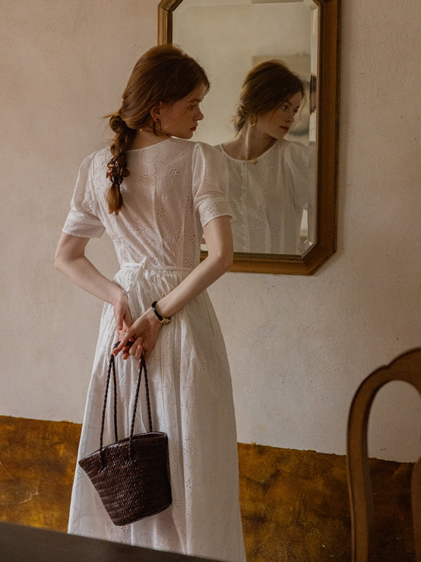 Back view of a woman holding a woven bag, styled in a white eyelet lace dress tied at the waist, standing in front of a mirror. Cottagecore aesthetic.