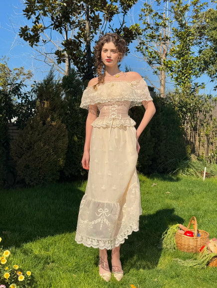 Full-length view of a woman wearing a vintage ivory lace corset dress with off-shoulder ruffle details, styled in a fairytale garden scene – romantic aesthetic women's outfit