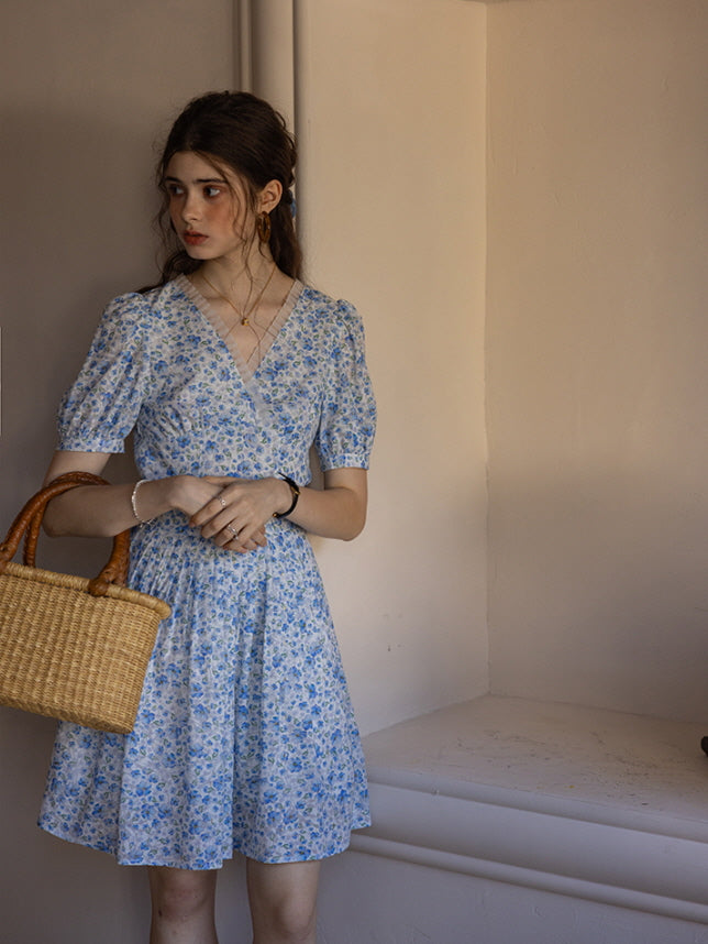 A woman in a short blue floral print dress with puff sleeves stands by a cream-colored wall, holding a woven basket. The setting exudes a soft, cottagecore aesthetic with delicate, vintage-inspired decor