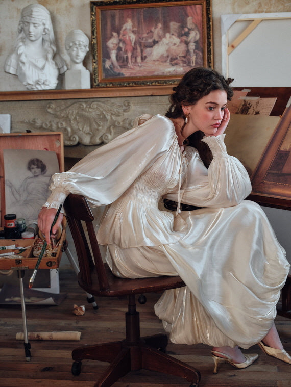 Woman in a romantic vintage ivory blouse and skirt, seated thoughtfully in an artist’s studio with classic statues and sketches in the background – elegant aesthetic fashion look.