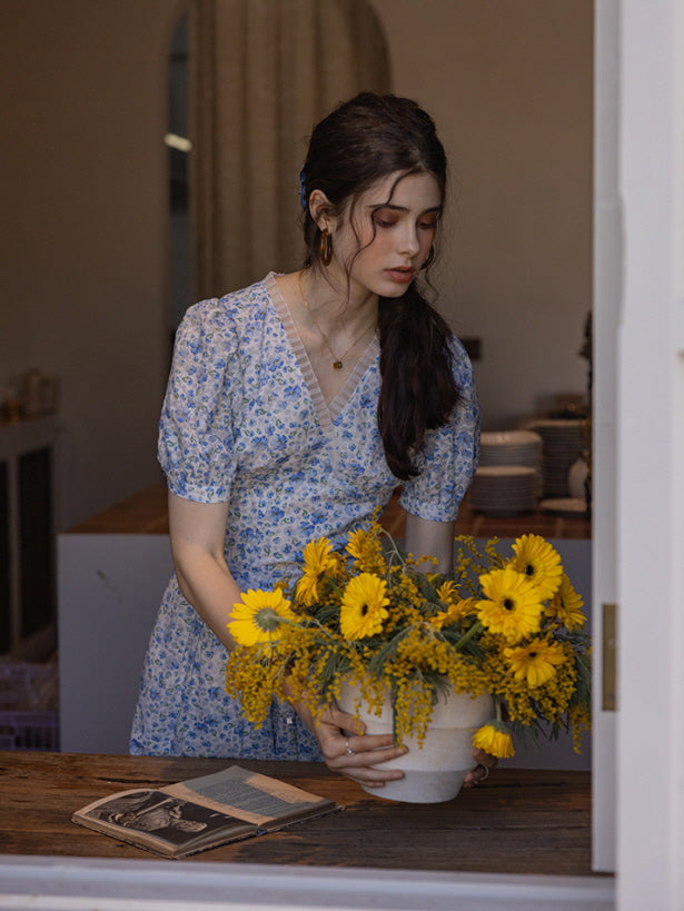 A woman in a short blue floral dress arranges a bouquet of vibrant yellow flowers in a white vase. The scene is set in a rustic kitchen, with a vintage book lying open on the wooden counter