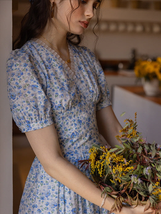 Close-up of a woman holding a bouquet of dried flowers while wearing a blue floral print dress with puff sleeves. The soft, vintage-inspired kitchen setting adds a cozy, cottagecore atmosphere.