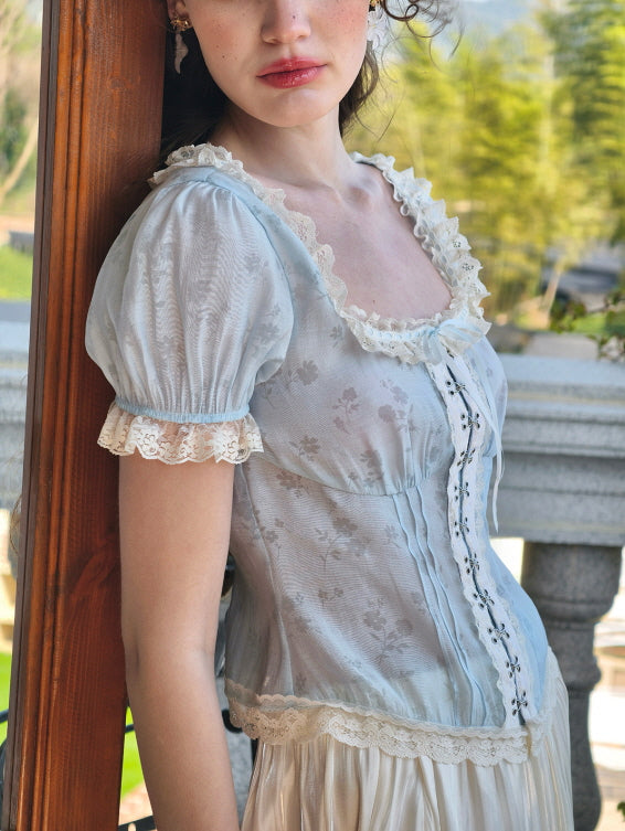 The model leans gracefully against a wooden frame, wearing a pastel blue corset top adorned with floral patterns and lace trim, exuding an ethereal, romantic aura amidst a serene garden backdrop at Romantic Fairytale store.