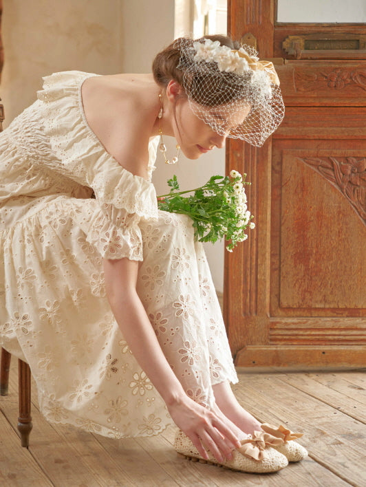 A woman in an off-shoulder embroidered dress adjusts her shoes in a vintage room, evoking a romantic cottagecore and fairytale mood.