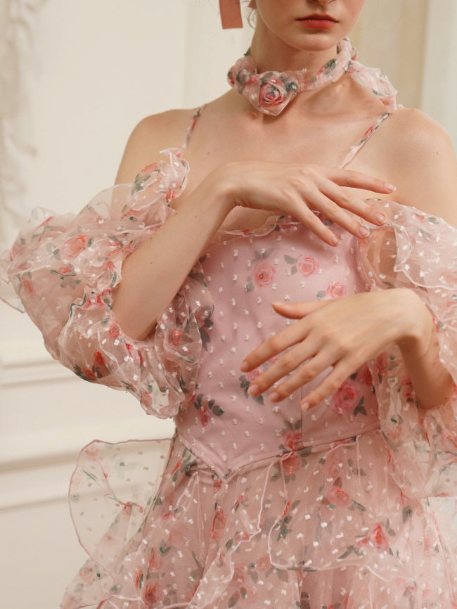 A woman wears a delicate pink floral chiffon dress with sheer puff sleeves and a rose choker, styled in a romantic fairycore aesthetic.