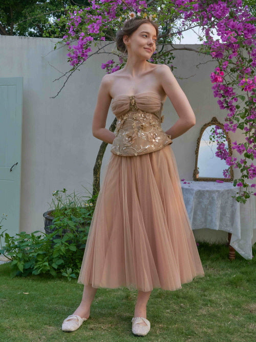 A woman stands in a blooming garden wearing a strapless tulle dress with embroidered flowers, evoking a soft fairycore and princesscore vibe.