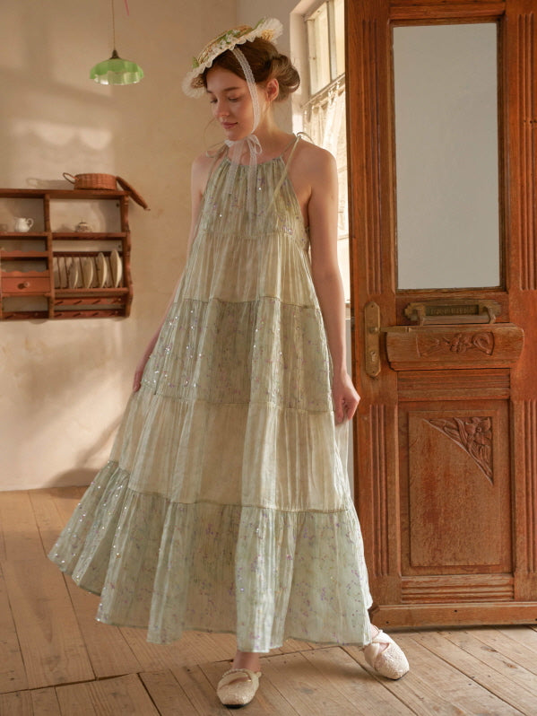 Wearing a long tiered floral dress with a soft shimmer, a woman stands in a rustic room styled with romantic cottagecore elegance.