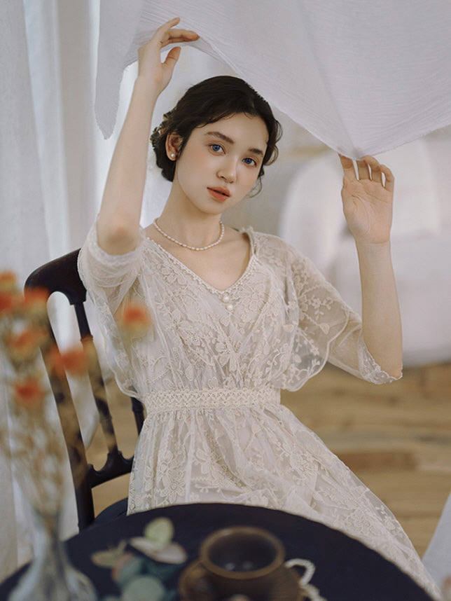 A young woman in a vintage-inspired lace dress sits gracefully, lifting a white curtain in a dreamy, cottagecore setting with soft lighting.