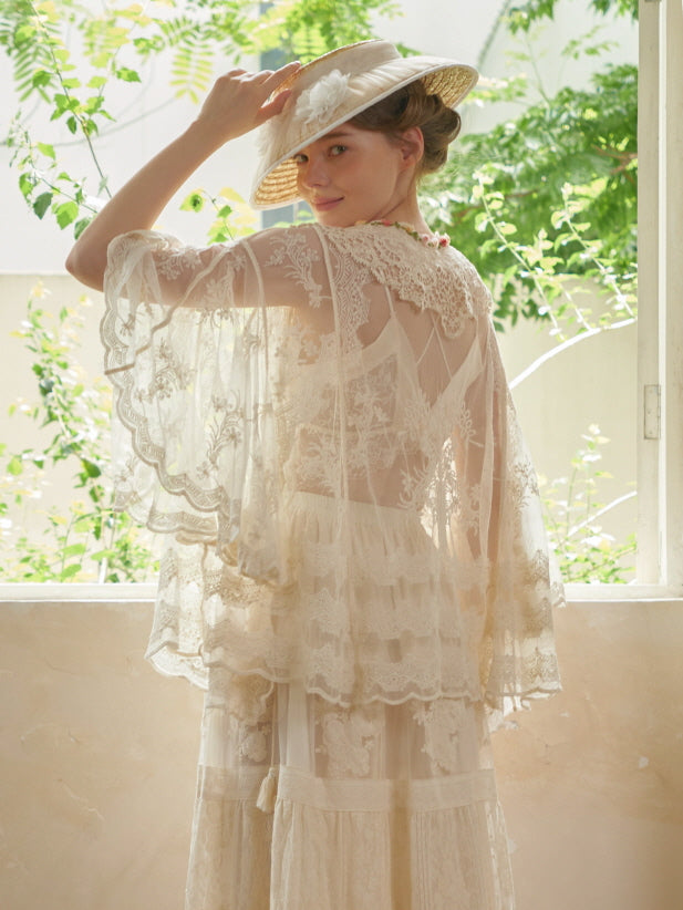 The model glances back with a soft smile, wearing a sheer lace cape and vintage skirt in a romantic fairycore setting.
