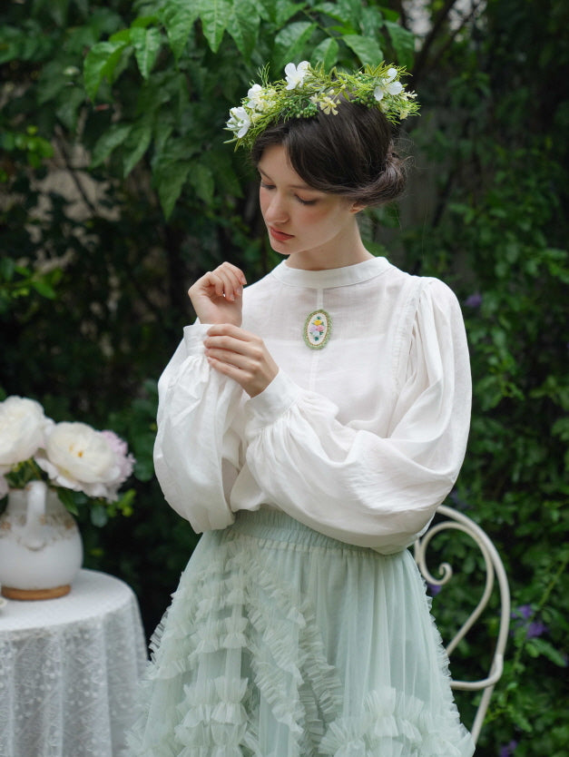 Wearing a puff-sleeve white blouse and ruffled pastel skirt, the model poses in a garden setting with a flower crown for a dreamy cottagecore look.